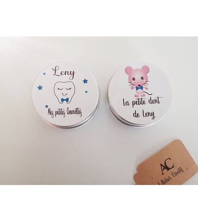 May include: Two small round metal tins with white lids. The tin on the left has a blue tooth with a bow tie and stars. The text on the tin reads "Leny" and "Mes petites quenottes". The tin on the right has a pink mouse with a blue bow tie. The text on the tin reads "La petite dent de Leny".