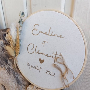 Peut inclure: Un cercle de broderie rond blanc avec le texte "Eveline et Clement" et un symbole de cœur. La date "16 juillet 2022" est écrite sous les noms.