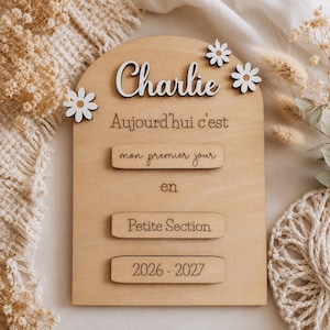 Peut inclure: Une plaque en bois en forme d'arche avec le prénom Charlie en relief blanc, trois fleurs blanches, et le texte gravé Aujourd'hui c'est mon premier jour en Petite Section 2026 - 2027 sur des éléments en bois horizontaux.