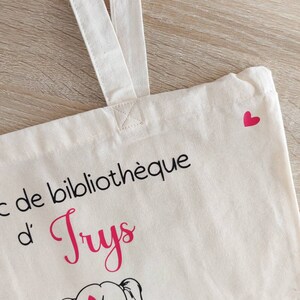 May include: A white canvas tote bag with black text that reads "Sac de biblioth&egrave;que d'Truys". A pink heart is in the top right corner of the bag. A cartoon illustration of a girl with pigtails reading a book is below the text. The book has the text "Truys &agrave; l'&eacute;cole" on the cover.
