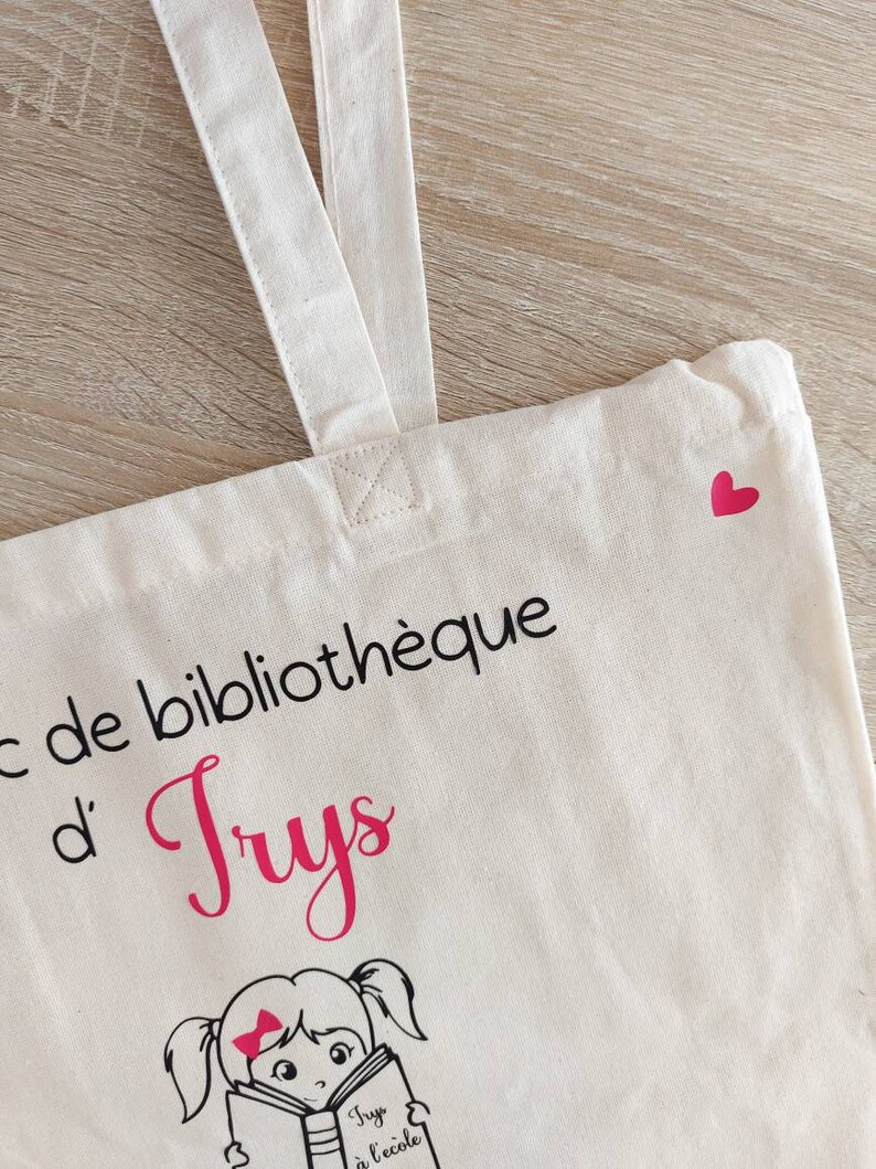 May include: A white canvas tote bag with black text that reads "Sac de biblioth&egrave;que d'Truys". A pink heart is in the top right corner of the bag. A cartoon illustration of a girl with pigtails reading a book is below the text. The book has the text "Truys &agrave; l'&eacute;cole" on the cover.