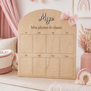 May include: Wooden photo display board with the name "Mya" in cursive. The board has spaces for school photos, labeled with French school grade abbreviations. A pink bow is attached to the top right.