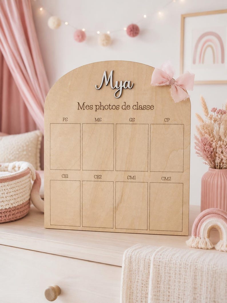 May include: Wooden photo display board with the name "Mya" in cursive. The board has spaces for school photos, labeled with French school grade abbreviations. A pink bow is attached to the top right.
