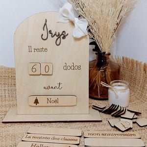 May include: Wooden countdown calendar with the name "Jeys" and the text "Il reste 60 dodos avant Noël". The calendar has a wooden base and a set of interchangeable wooden tiles with various events, including "Halloween", "La rentrée des classes", "Le départ en vacances", "mon anniversaire", and "Pâques".