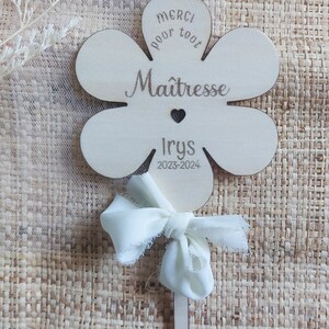 May include: A wooden flower-shaped sign with the text "Merci pour tout Ma&icirc;tresse Irys 2023-2024". The sign is attached to a stick with a white ribbon bow.