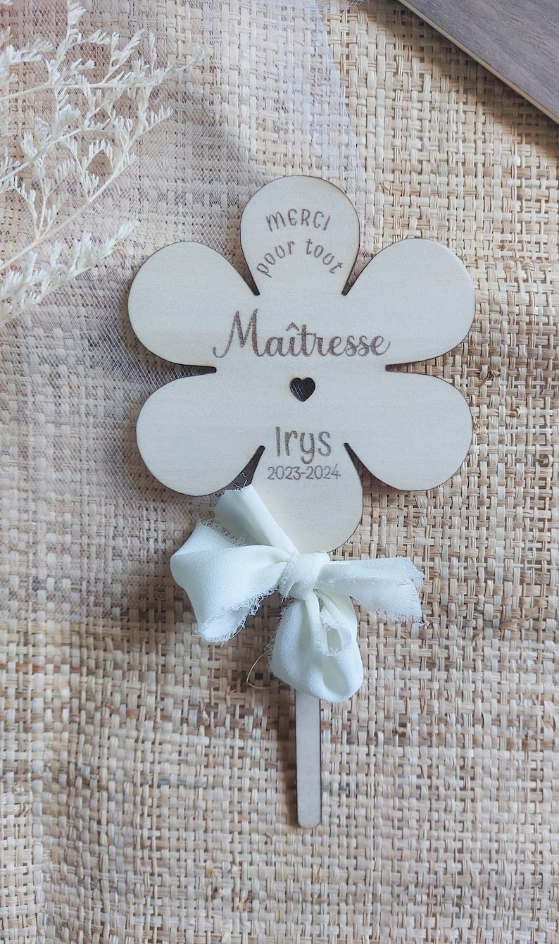 May include: A wooden flower-shaped sign with the text "Merci pour tout Ma&icirc;tresse Irys 2023-2024". The sign is attached to a stick with a white ribbon bow.