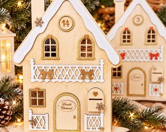 May include: A light wood advent calendar house featuring a white roof, gold glitter accents, and a central door labelled Famille Chabanel. The structure includes a balcony, windows, a mailbox, and a circular number 14 disc on the front facade.