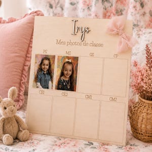 May include: A wooden photo display board with the name "Irys" and the text "Mes photos de classe". The board has spaces for school photos, with two photos displayed. A pink bow is attached to the top right corner. A stuffed bunny sits in the lower left.