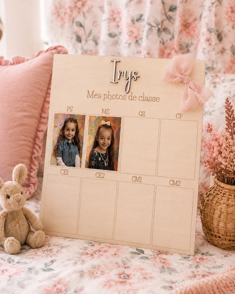 May include: A wooden photo display board with the name "Irys" and the text "Mes photos de classe". The board has spaces for school photos, with two photos displayed. A pink bow is attached to the top right corner. A stuffed bunny sits in the lower left.