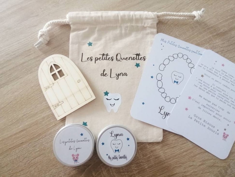 May include: A tooth fairy kit containing a wooden door, a tooth-shaped box, and a small jar of tooth-friendly treats. The kit is decorated with stars and the text "Les petites Quenottes de Lyna".