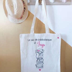 May include: White canvas tote bag with pink hearts and a black and white illustration of a girl reading a book. The text on the bag reads "Le sac de biblioth&egrave;que d'Tryss".
