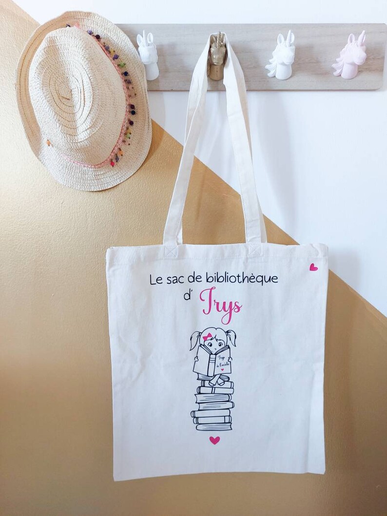 May include: White canvas tote bag with pink hearts and a black and white illustration of a girl reading a book. The text on the bag reads "Le sac de biblioth&egrave;que d'Tryss".