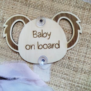 May include: A wooden "Baby on board" sign shaped like a koala's head. The sign is light brown with darker brown detailing and features two suction cups. The words "Baby on board" are engraved on the sign.