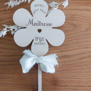 May include: A wooden flower-shaped sign with the text "Merci pour tout Ma&icirc;tresse Iry's 2023-2024" and a small heart. The sign is attached to a wooden stick with a white fabric bow.