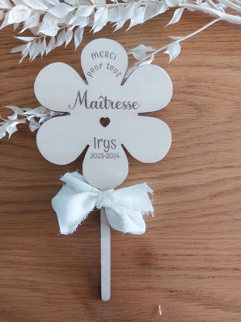 May include: A wooden flower-shaped sign with the text "Merci pour tout Ma&icirc;tresse Iry's 2023-2024" and a small heart. The sign is attached to a wooden stick with a white fabric bow.