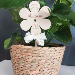 May include: A wooden flower-shaped sign with the text "Merci pour tout Ma&icirc;tresse Iry's 2023-2024" in brown lettering. The sign is attached to a plant in a woven basket.