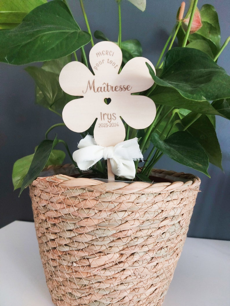 May include: A wooden flower-shaped sign with the text "Merci pour tout Ma&icirc;tresse Iry's 2023-2024" in brown lettering. The sign is attached to a plant in a woven basket.