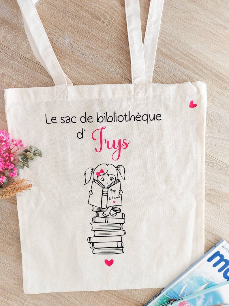 May include: A white canvas tote bag with pink hearts and black text that reads "Le sac de biblioth&egrave;que d'Trys". A black and white illustration of a girl sitting on a stack of books and reading a book that says "J'app &agrave; l'&eacute;cole" is on the bag.
