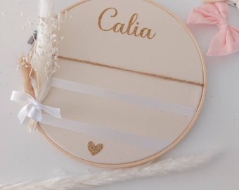 May include: A white embroidery hoop with the name "Calia" written in gold glitter. The hoop has two white ribbons and a gold glitter heart in the center. There are dried white pampas grasses on either side of the hoop.