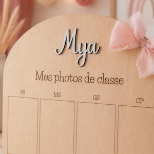 May include: Wooden photo display board with the name "Mya" in silver script. The board has spaces for school photos and the text "Mes photos de classe". A pink bow adorns the top right corner.