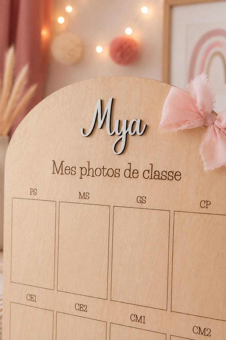 May include: Wooden photo display board with the name "Mya" in silver script. The board has spaces for school photos and the text "Mes photos de classe". A pink bow adorns the top right corner.
