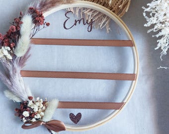 May include: A round wooden hoop with a brown mesh fabric stretched across it. The hoop is decorated with dried flowers and a brown ribbon. The name "Emy" is written in cursive on the hoop. A small heart with glitter is also on the hoop.