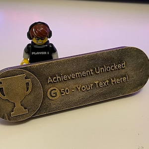 May include: A gold-coloured plastic plaque with a trophy icon and the text "Achievement Unlocked G 50 - Your Text Here!"
