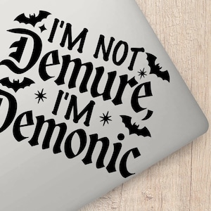 May include: Black and white sticker with the text "I'm not demure I'm demonic" in a gothic font. The sticker features bats and stars.