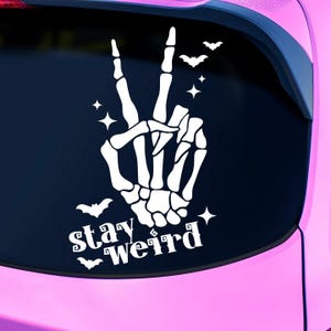 Op de afbeelding: Een witte skelethand die een peace-teken maakt met de tekst "stay weird" eronder. De achtergrond is zwart met witte vleermuizen en sterren.