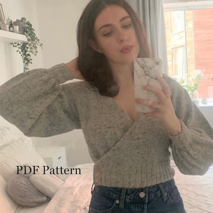 Puede incluir: Un suéter gris de punto con cuello en V y mangas largas. El suéter tiene un patrón moteado y es corto a la cintura. La modelo lleva el suéter con jeans azules y tiene un teléfono en la mano. El texto "PDF Pattern" es visible en la imagen.
