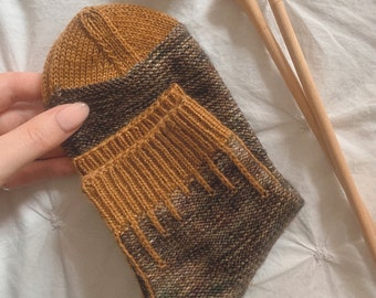 Może przedstawiać: Para skarpet knitted w kolorze brązowym i musztardowym z kontrastowym wzorem. Skarpety są złożone i trzymane w dłoni. W tle znajdują się dwa drewniane druty do robótek na drutach.
