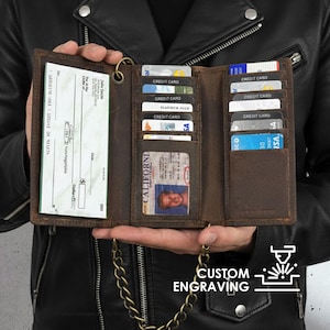 Può includere: Un portafoglio in pelle marrone con più fessure per carte, un portabiglietti da visita e una finestra di identificazione trasparente. Il portafoglio è aperto, mostrando carte di credito, un documento d'identità della California e un assegno. È attaccata una catena di metallo. Il testo "CUSTOM ENGRAVING" è visibile.