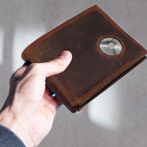 May include: A brown leather wallet with orange stitching and a silver circular accent. The wallet is held in a hand against a gray background. The leather has a distressed appearance.