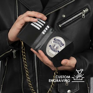 May include: Black leather badge holder with a gold chain, holding a silver security officer badge. The badge has the words "SECURITY ENFORCEMENT OFFICER" in a blue and silver design. The holder has slots for cards. The image also includes the text "CUSTOM ENGRAVING".