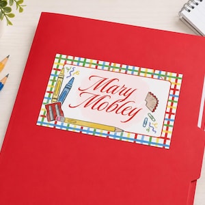 May include: A bright red file folder featuring a rectangular label with a colorful plaid border. The label displays the name Mary Mobley in red script, surrounded by illustrations of a yellow pencil, a blue crayon, a sharpener, and paper clips.