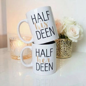Könnte beinhalten: Zwei weiße Keramikbecher, die übereinander gestapelt sind. Der obere Becher trägt die Aufschrift "HALF his DEEN" in schwarzer und goldener Schrift. Der untere Becher trägt die Aufschrift "HALF her DEEN". Eine Kerze und Blumen sind im Hintergrund.