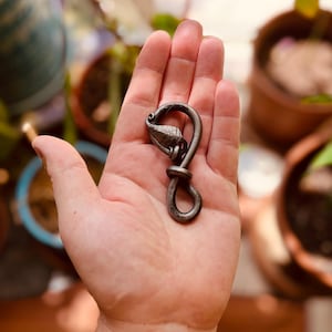 May include: A hand-forged metal keychain with a unique, artistic design. The keychain features a loop at the bottom, a knot in the middle, and a stylized leaf or petal shape at the top. The metal has a dark, aged finish.