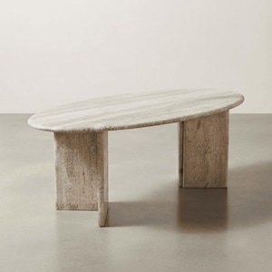 May include: An oval coffee table crafted from beige travertine stone with a natural porous texture. The table features a smooth, elongated top surface supported by two thick, rectangular slab legs that provide a sturdy and balanced base structure.