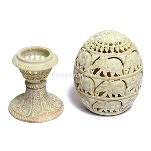 Puede incluir: Dos objetos decorativos de piedra tallada. Uno es un pedestal con un diseño floral. El otro es un recipiente en forma de huevo con tallas de elefantes y detalles calados. Ambos son de color beige claro.