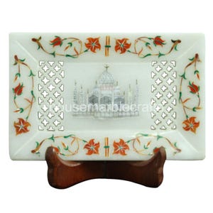 Könnte beinhalten: Ein rechteckiges Tablett aus weißem Marmor mit einer detaillierten Taj Mahal-Illustration in der Mitte. Das Tablett hat einen floralen Rand mit roten und orangen Akzenten und wird von einem Holzständer getragen. Das Tablett hat ein dekoratives Ausschnittdesign.