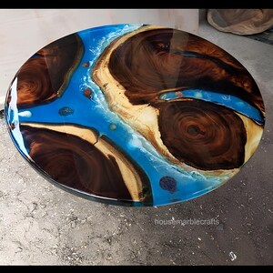 Op de afbeelding: Een rond tafelblad met natuurlijke donkerbruine houten platen met opvallende nerven en natuurlijke randen, gescheiden door een glanzende, doorschijnende blauwe harsinleg met witte wervelende details. De tekst housemarblecrafts is zichtbaar.