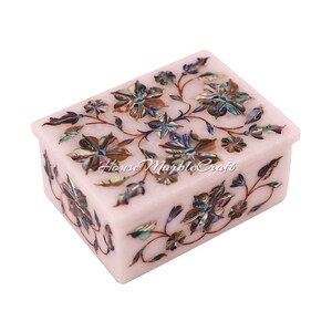 May include: A rectangular white marble box with floral inlay. The box features a lid and is adorned with a detailed floral pattern in shades of brown, blue, and iridescent shell. The design is symmetrical, with flowers and leaves.