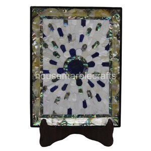 May include: Rectangular decorative marble piece with a central sunburst design in white, blue, and iridescent shell accents. The piece is framed in black and rests on a dark brown wooden stand. The text "housemarblecrafts" is visible.