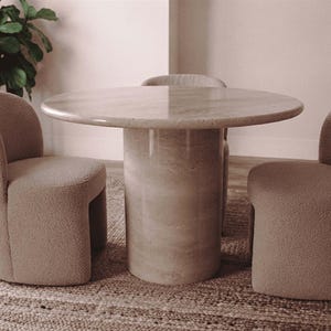 May include: A round dining table with a smooth, light beige stone top and a cylindrical pedestal base, accompanied by three upholstered chairs featuring a textured, light tan fabric and curved backrests arranged around the table.