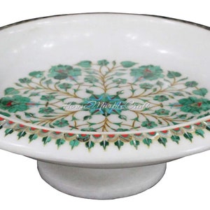 May include: White marble decorative dish with a pedestal base. The dish features a floral inlay design in shades of green, gold, and blue. The rim is adorned with a decorative border. The text "Home Marble Craft" is visible.