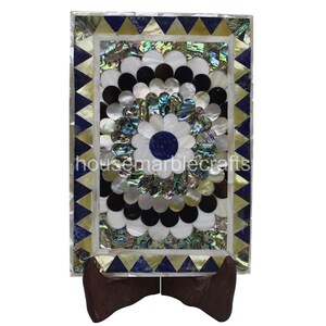 May include: A rectangular decorative marble piece with a floral design. The design features white, black, and iridescent shell-like elements, with a blue center. The border is adorned with blue and gold triangles. It is displayed on a wooden stand.