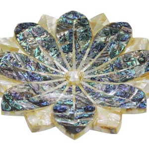 Op de afbeelding: Een decoratieve, bloemvormige onderzetter met een ontwerp van parelmoer en abalone schelp. De onderzetter heeft iriserende blauwe, groene en zilveren tinten, met een crèmekleurige buitenrand. De bloemblaadjes zijn in een symmetrisch patroon gerangschikt.