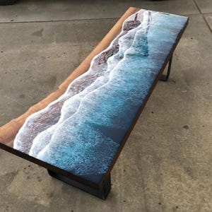 May include: A long wooden bench with a blue and white epoxy resin top that resembles ocean waves. The bench has black metal legs.
