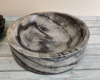 May include: A circular, grey marble bowl with a swirling pattern of black and white. The bowl has a wide, shallow design and the text "House Marble Craft" is visible on the inside. The bowl is on a light blue wooden surface.