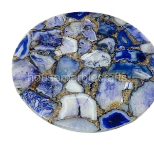 May include: Circular stone coaster with a mosaic pattern. It features blue and white stone pieces set within a gold-coloured matrix. The surface is polished and reflective, suitable for drinks.